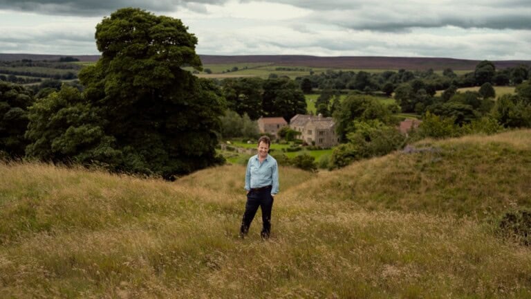 David Hill stood in a meadow with his Yorkshire home in the background