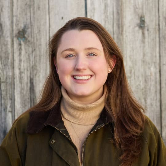 Elizabeth is a woman in her twenties with long, straight brown hair wearing a beige turtle-neck jumper and green waxed countryside jacket