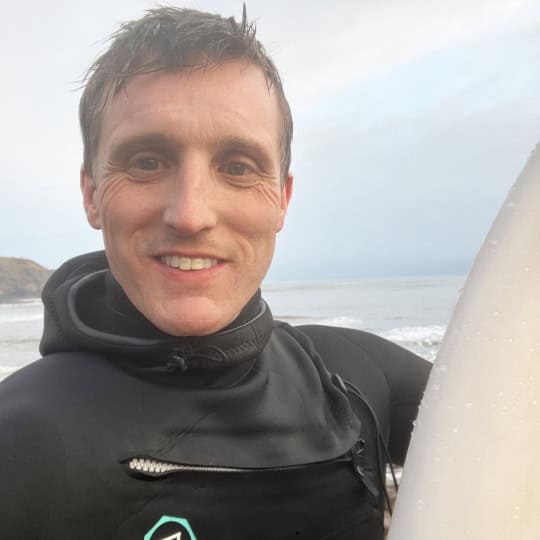 Fraser is a man in his forties with short brown hair wearing a black wetsuit, holding a surfboard
