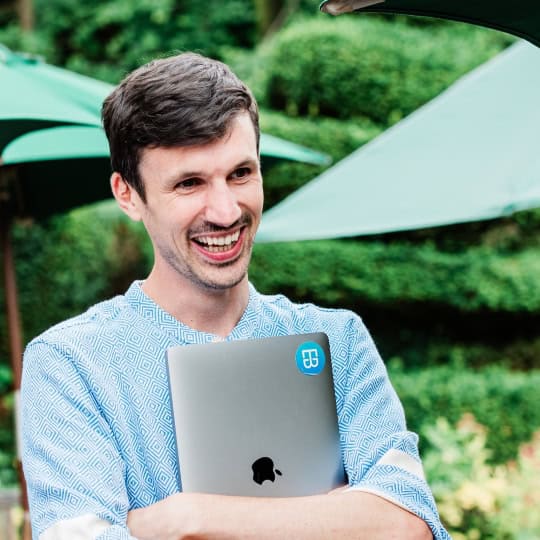 George is a man in his thirties with short, dark brown hair and stubble wearing a blue shirt, holding a laptop