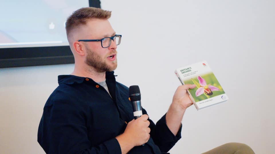 Environment Bank ecologist, Mike Waller, a man in his thirties with a button-up blue shirt, glasses, and short brown hair, speaking at an event, holding a copy of his book on Britain's orchids
