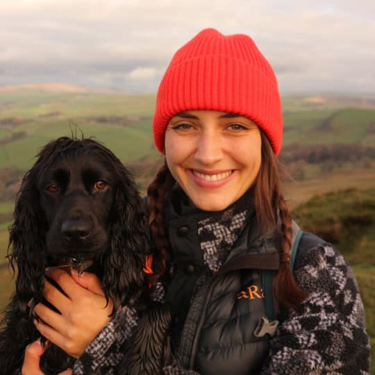 Nikita is a woman in her twenties with shoulder-length brown hair wearing a bright red beanie hat and hiking jacket holding her dog, a black spaniel called Pip