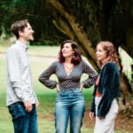 Three Environment Bank employees casually dressed stood chatting at summer party; one a woman in her twenties with curly red hair, one a woman in her thirties with shoulder-length black hair with dyed purple ends, and one a man in his thirties with short brown hair and stubble