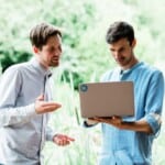 Two Environment Bank employees looking at laptop screen together, both men in their thirties wearing button-up shirts