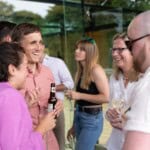 Four Environment Bank employees dressed casually, chatting and laughing together with drinks at summer party, men and women between the ages of twenty and sixty