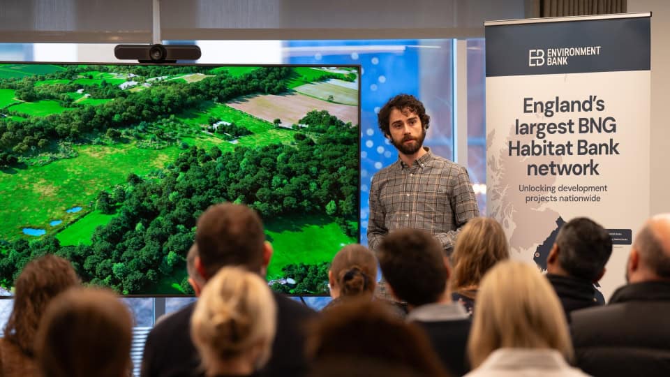 Environment Bank ecologist speaking at a BNG Breakfast event, a man in his thirties with curly black hair and beard, stood beside a large screen showing a bright birds-eye vision of one of our Habitat Banks