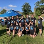 Twelve Environment Bank employees wearing matching navy t-shirts and shorts, some with sunglasses and hats, all between the ages of twenty and fifty, the team for the 2023 charity walk