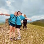 Two Environment Bank employees on a company charity walk wearing blue t-shirts, black shorts, and hiking trainers - one, a woman with short blonde hair and the other, a non-binary person in their thirties with several leg and arm tattoos wearing glasses and a backwards baseball cap