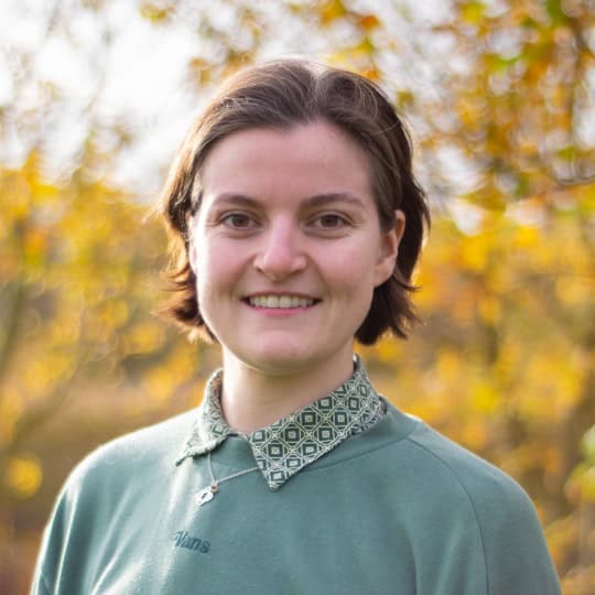 Ell is a non-binary person in their early thirties with short brown hair wearing a green button-up shirt and green jumper