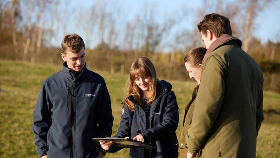 Four members of the Environment Bank team at a Habitat Bank, two wearing waxed country jackets and two wearing blue zip-up jackets, gathered around a clipboard - two are men in their thirties and two are women in their twenties