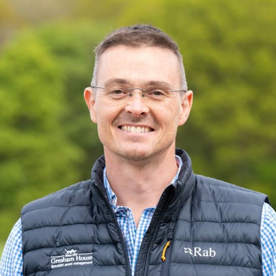 Chris is a man in his fifties with short brown hair wearing rimless glasses, a check blue button-up shirt, and a navy, Gresham House-branded bodywarmer
