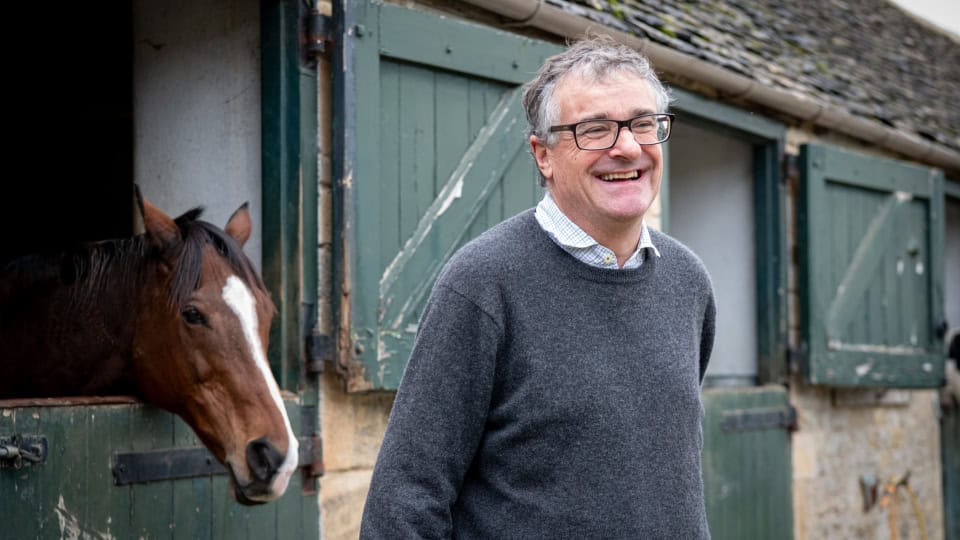 Alex, landowner and estate manager at Cornwell Habitat Bank, a man in his sixties with grey hair and glasses wearing a grey wool jumper stood in front of stables with a brown horse stood behind him