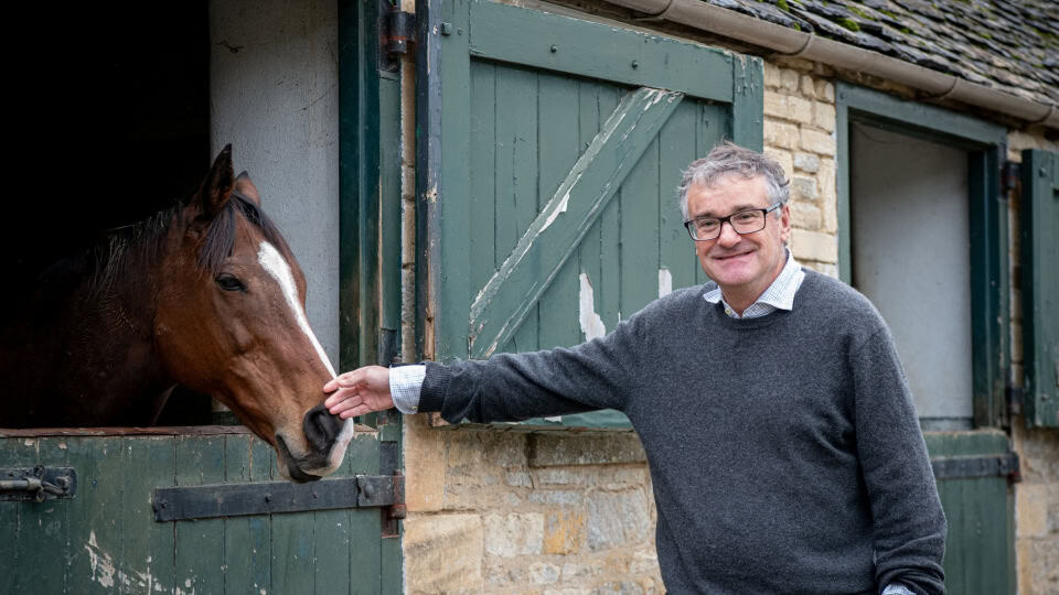 Alex, landowner and estate manager at Cornwell Habitat Bank, a man in his sixties with grey hair and glasses wearing a grey wool jumper stood next to stables, petting a brown horse