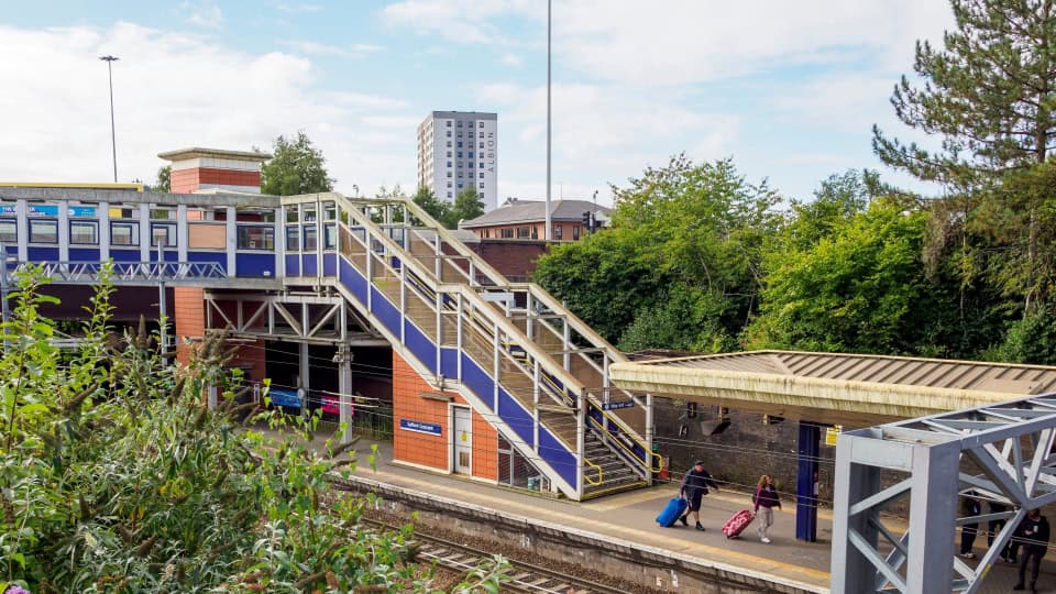Looking down onto Salford Crescent Station with a bridge crossing the railway tracks, passengers carrying suitcases, and trees and bushes surrounding the station