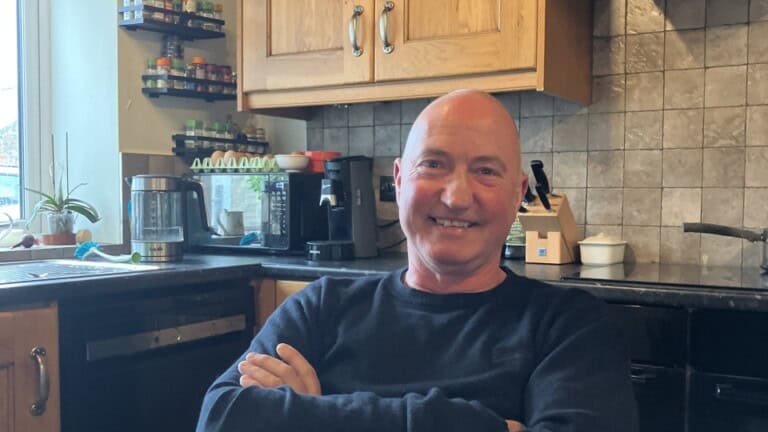 Richard Routledge, Environment Bank's BNG farmer partner at Heighington Habitat Bank, a man in his fifties wearing a blue jumper, sat in his kitchen, smiling