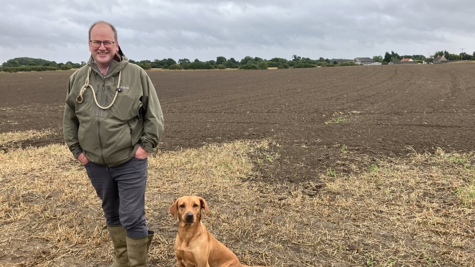 Environment Bank's land manager, Will Witchell, a man in his fifties wearing a green anorak, grey trousers, and wellington boots, stood at Heighington Habitat Bank in front a field prepared for new grasslands, with his dog