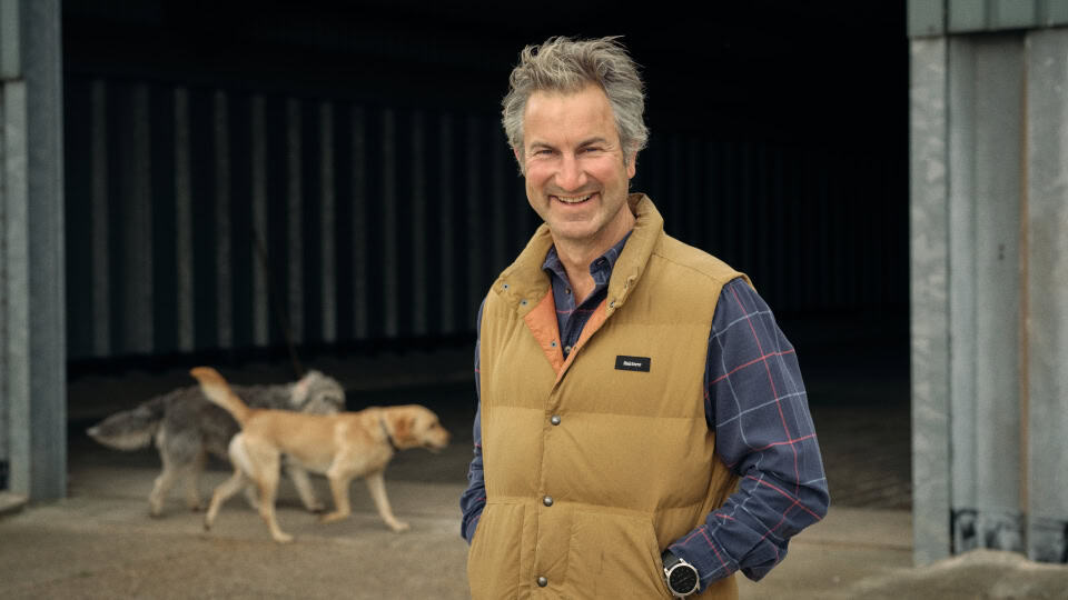 Environment Bank's landowner partner, James Bucher, a man in his forties with grey and brown hair wearing a check blue shirt and mustard gilet - stood in front of a farm steading where James' dogs are walking