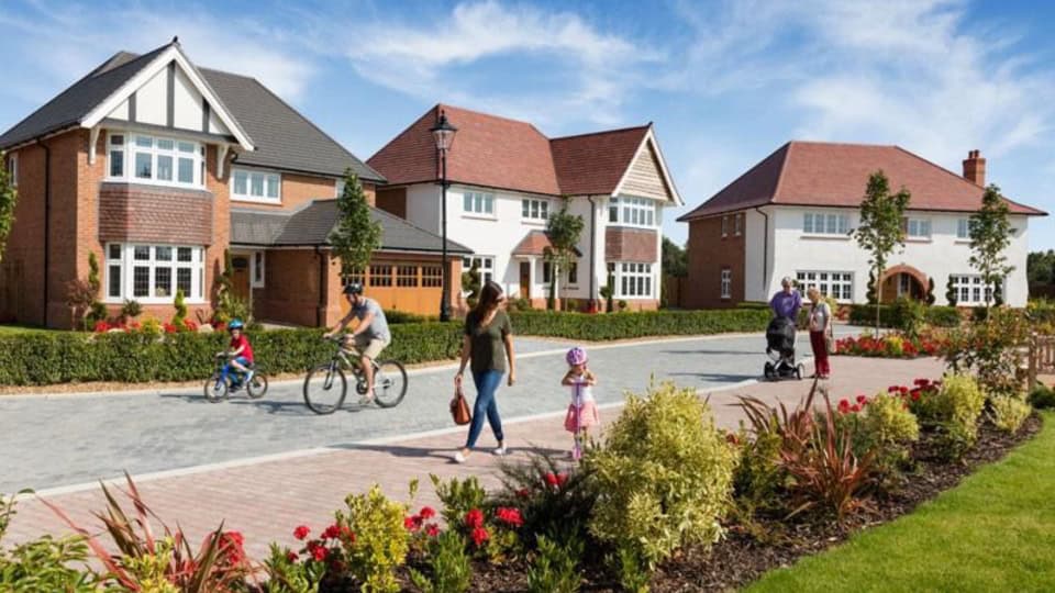 Visualisation of Redrow's new development showing a peaceful residential street with three large detached houses, lots of hedges, bushes, flowers, small trees, and a grassy area. Residents depicted on the footpath and street, including a young child and adult riding their bikes, an adult walking with a small child riding a scooter, and a pair of people stood talking while one holds a buggy.