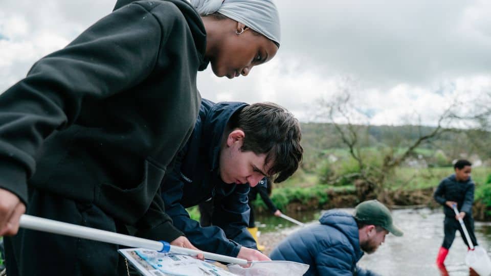 Two young people from Black2Nature are standing in a river, looking into a fishing net.